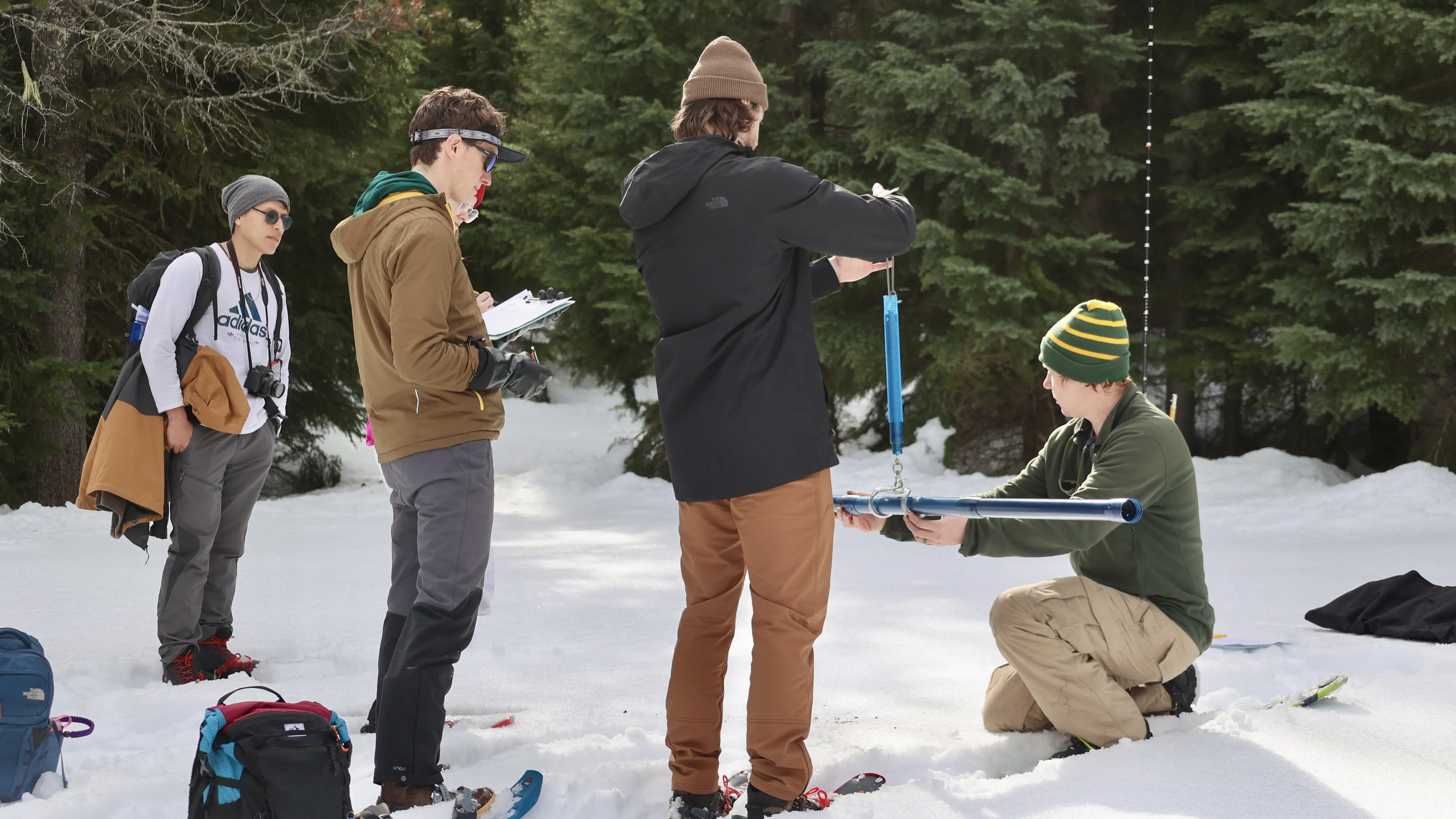 A researcher kneels in the snow with a measuring device as students with notebooks look on. 