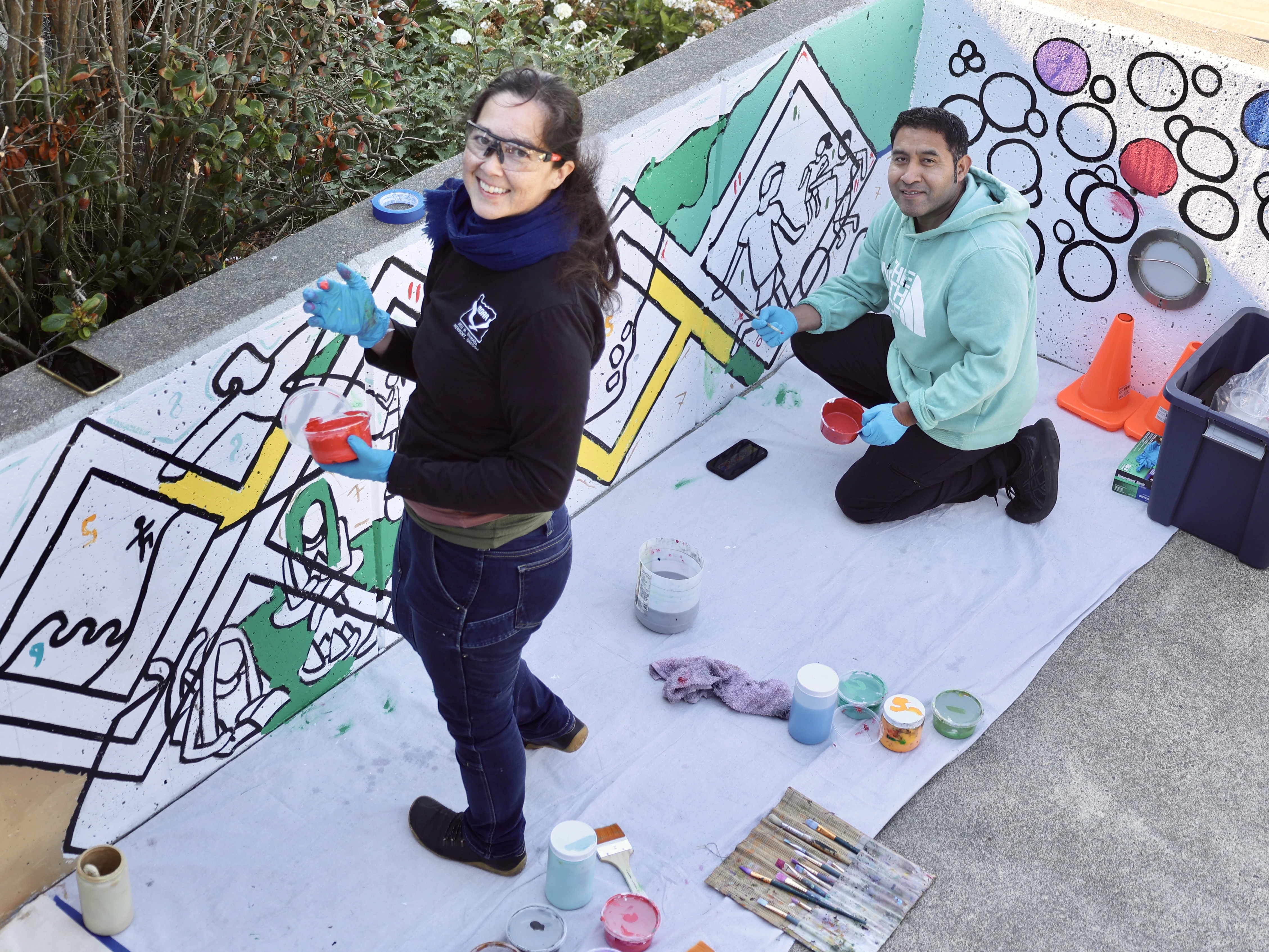 Two artists look up smiling from mural in progress.